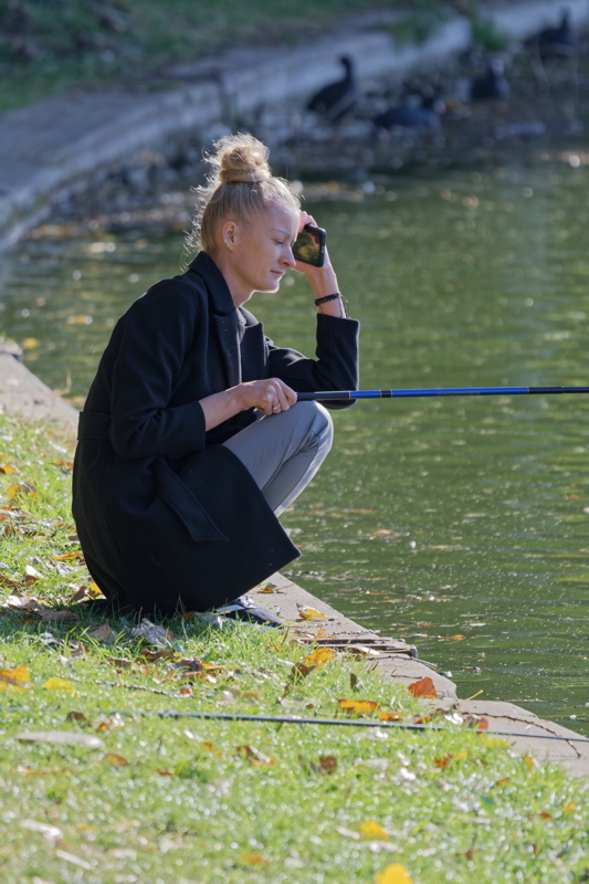14 - A woman squatting by the edge of a body of water, holding a fishing rod while talking on a mobile phone